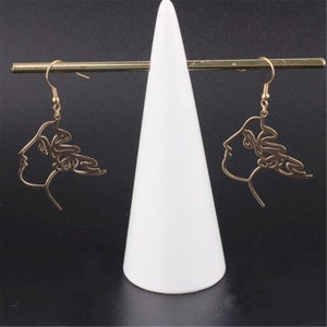 May include: A pair of gold-toned earrings with a line drawing design of a woman's face. The earrings are hanging from a white display stand.