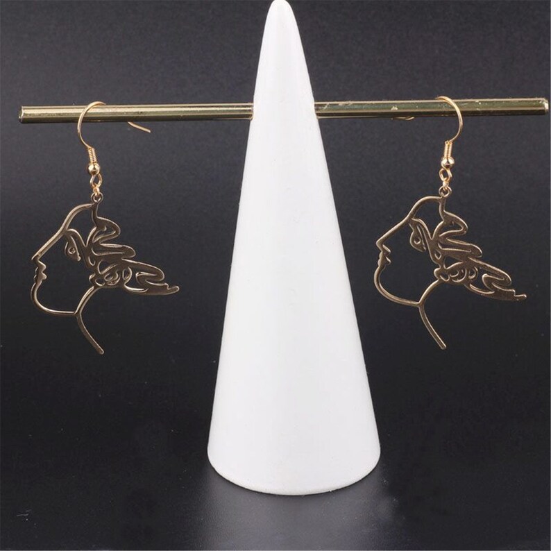 May include: A pair of gold-toned earrings with a line drawing design of a woman's face. The earrings are hanging from a white display stand.