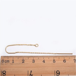 May include: A gold chain earring with a curved hook and a straight bar extending from the chain. The earring is measured on a ruler, showing it is approximately 7 cm long.