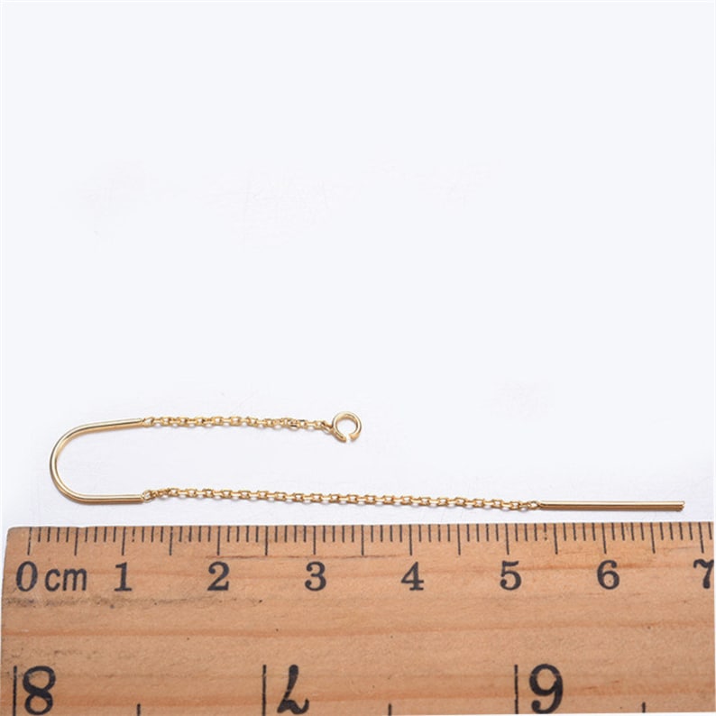May include: A gold chain earring with a curved hook and a straight bar extending from the chain. The earring is measured on a ruler, showing it is approximately 7 cm long.