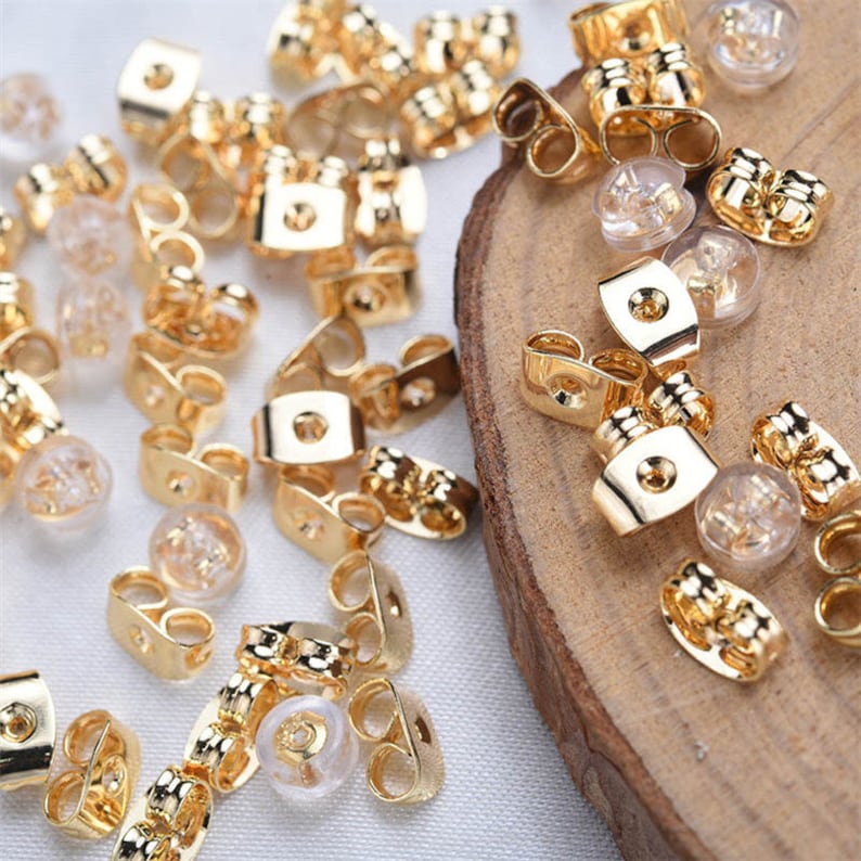 May include: A collection of gold-colored earring backs and clear, rounded earring stoppers are scattered on a wooden surface. The earring backs have a rectangular shape with a small hole in the center. The stoppers are made of a clear, rubbery material.