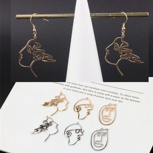 May include: A pair of gold-toned earrings with a line art design of a woman's face. The earrings are shown next to five additional line art designs of faces, two in gold and three in silver.