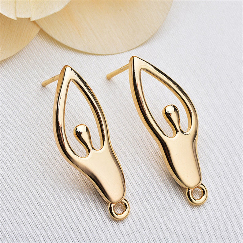 May include: A pair of gold-tone earrings featuring a stylized human figure design. The earrings have a loop at the bottom for attaching to an earring post.