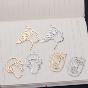 May include: Six gold and silver metal charms in the shape of abstract faces. The charms are outlined and have a hole at the top for hanging.