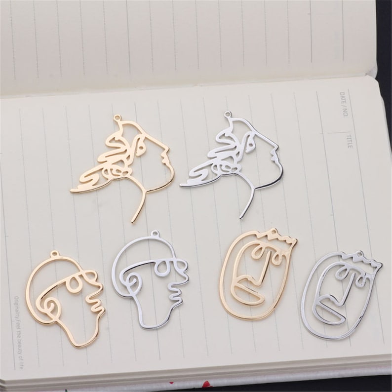 May include: Six gold and silver metal charms in the shape of abstract faces. The charms are outlined and have a hole at the top for hanging.
