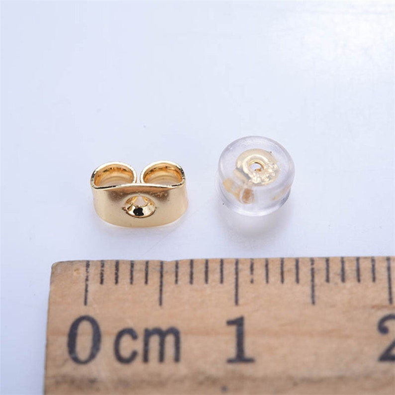 May include: Two earring backs are shown. One is a gold-colored butterfly-shaped earring back. The other is a clear, round earring back with a gold-colored metal insert. A wooden ruler with centimeter markings is in the foreground.