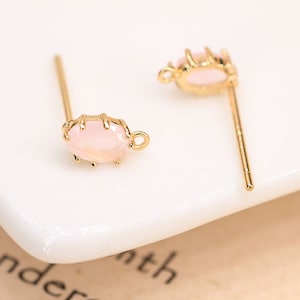 May include: A pair of gold-tone earring posts with a pink oval-shaped stone set in a gold-tone setting.