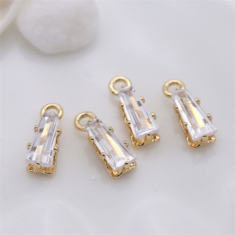 May include: Four gold-toned pendant charms with clear rectangular crystal stones. Each charm has a loop at the top for attaching to a necklace or bracelet.