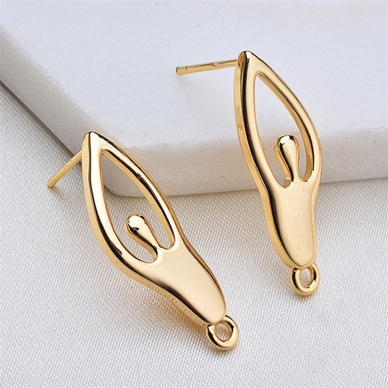 May include: A pair of gold-tone earrings with a unique design featuring a stylized human figure. The earrings have a post back closure.