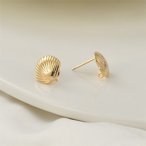 May include: A pair of gold-colored seashell stud earrings. The earrings are designed with a detailed, ridged texture, mimicking the shape of a scallop shell. The posts are thin and straight, with a small loop at the end. The earrings are displayed on a white surface.