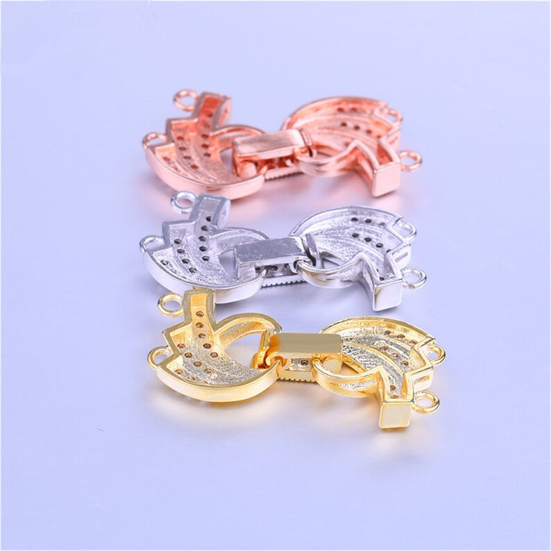 May include: Three different colored metal clasps for necklaces or bracelets. The clasps are in the shape of a crescent moon with a decorative design and have a small, rectangular latch. The clasps are gold, silver, and rose gold.