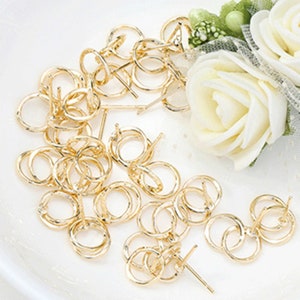 May include: A close-up of a pile of gold-toned earring findings. The findings are made of metal and have a small loop at the top and a post at the bottom. The findings are arranged in a random pattern on a white surface.