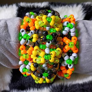 May include: A wide cuff bracelet constructed from numerous plastic pony beads in a vibrant color palette of neon green, bright orange, sunny yellow, opaque white, and translucent black, arranged in a complex, multi-layered geometric pattern.