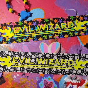 May include: Two fabric headbands with the text "EVIL WIZARD". One headband has a black background with colorful stars, the other has a black background with white skulls and bones. Both have yellow flames.