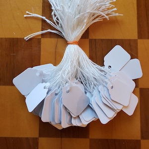 May include: A bundle of white string with a bunch of small, white, rectangular gift tags attached. The tags have a small hole punched in the top and are tied together with a rubber band.