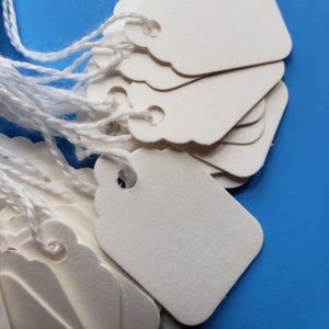 May include: A stack of white paper gift tags with string attached. The tags have scalloped edges and a hole punched in the top.