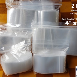 May include: A large quantity of clear plastic resealable bags, 2 mil thick, 4 inches by 4 inches, with a zipper closure. The bags are stacked and arranged in a pile on a wooden surface. The text "2 Mil", "Free Shipping", "2-3 Day Delivery", "Best Quality ~ Best Deal", and "4" x 4" is printed on a white label.