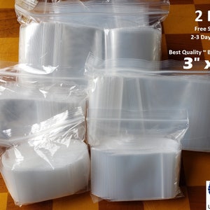 May include: A large stack of clear plastic resealable bags, 2 mil thick, 3 inches by 5 inches. The bags are arranged in a pile on a checkered surface. The text "2 Mil", "Free Shipping", "2-3 Day Delivery", "Best Quality Best Deal", and "3" x 5" is printed on a white label.