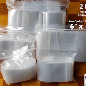 May include: A large quantity of clear plastic resealable bags, 2 mil thick, 6 inches by 10 inches. The bags are stacked and arranged on a wooden surface. The text "2 Mil", "Free Shipping", "2-3 Day Delivery", "Best Quality Best Deal", and "6" x 10" is printed on a white label.