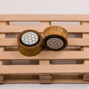 May include: Two wooden ear gauges with a flower of life design. The gauges are brown and have a white flower of life design on the front.