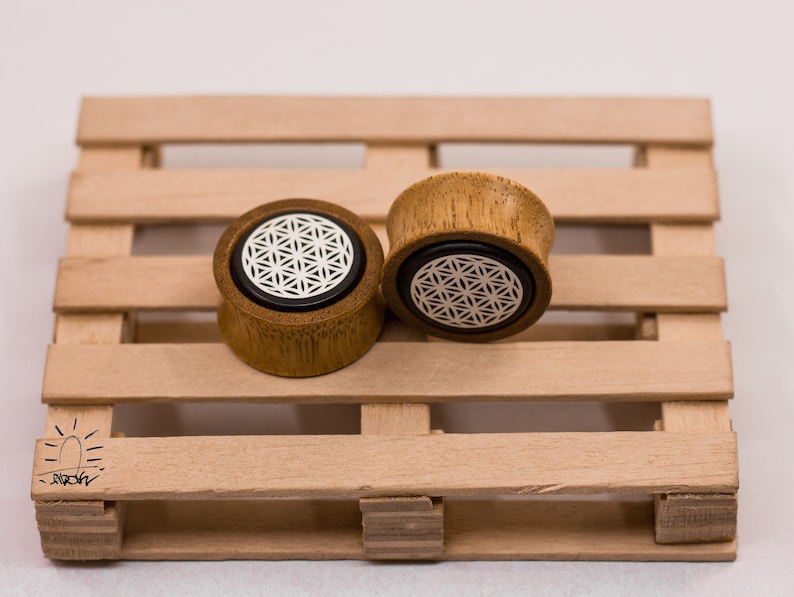 May include: Two wooden ear gauges with a flower of life design. The gauges are brown and have a white flower of life design on the front.