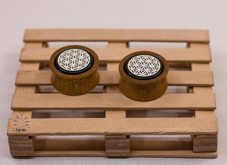 May include: A pair of wooden ear gauges with a black and white flower of life design. The gauges are on a wooden pallet.