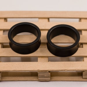 May include: Two black acrylic ear gauges, also known as plugs or tunnels, are displayed on a wooden pallet. The gauges are circular with a smooth, polished finish.