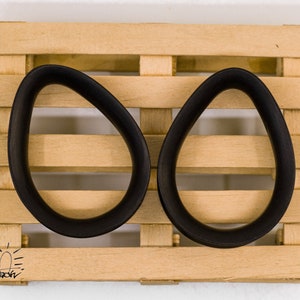 May include: Two black wooden ear gauges, shaped like ovals, are displayed on a wooden pallet. The gauges are smooth and have a glossy finish.