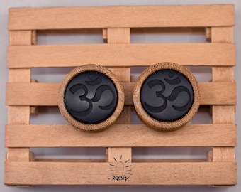 May include: Two round, wooden plugs with a black Om symbol in the center. The plugs are set against a light brown wooden pallet background. The Om symbol is a spiritual icon.