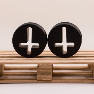 May include: Pair of black wooden ear gauges with an upside down cross cutout design. The gauges are displayed on a small wooden pallet.