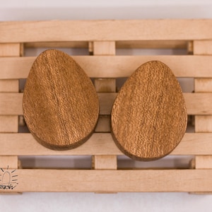 May include: Two brown wooden ear gauges, shaped like ovals, are displayed on a wooden pallet. The gauges are smooth and have a natural wood finish.