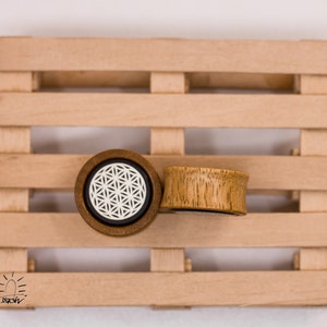 May include: Two wooden ear gauges, one with a white geometric pattern and the other with a natural wood finish, displayed on a wooden pallet.