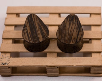 May include: Two teardrop-shaped wooden ear gauges with a dark brown wood grain pattern. The gauges are sitting on a wooden pallet.