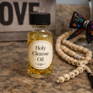 May include: A small glass bottle of Holy Cleanse Oil with a black cap. The bottle contains a golden liquid with small particles. The label is oval-shaped with the words "Holy Cleanse Oil" in a serif font. A wooden bead necklace and a decorative bow are in the background.