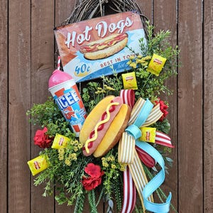May include: A decorative wreath featuring a vintage "Hot Dogs" sign, a plush hot dog, a pink and blue ice drink, and colorful ribbons. The wreath is adorned with greenery, red flowers, and small yellow snack bags, creating a festive summer theme.