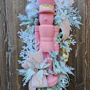 May include: A pink and gold Christmas wreath featuring a pink nutcracker, white and gold poinsettias, and a variety of greenery.