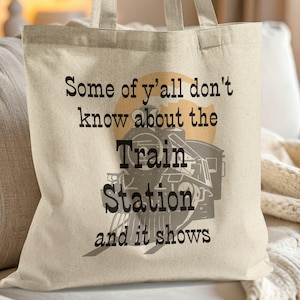May include: A beige canvas tote bag featuring a black graphic of a steam locomotive train engine centered over a tan circular background. The text reads: Some of y'all don't know about the Train Station and it shows, printed in black lettering.