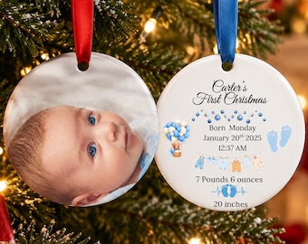 May include: Two round ceramic ornaments hanging by red and blue ribbons. One features a photograph of a baby. The other displays text: Carter's First Christmas, Born Monday January 20th 2025, 12:37 AM, 3345 grams, 51 cm, with blue baby footprints and a clothesline graphic.