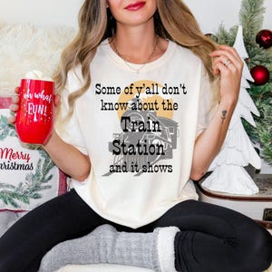 May include: Cream-colored t-shirt with a graphic of a train and the text "Some of y'all don't know about the Train Station and it shows." The shirt is paired with black leggings and gray socks. A red mug with "oh what FUN!" is held in the hand.