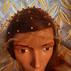 Puede incluir: Un velo vintage con un delicado tejido de red y pequeñas cuentas rosas. El velo se lleva en una cabeza de maniquí con cabello castaño.