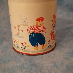 May include: A vintage metal canister with a red lid and a cream-colored body. The canister is decorated with Dutch folk art illustrations of people, windmills, and tulips. The canister is cylindrical in shape.