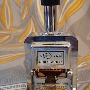 Puede incluir: Un sello de numeración automática de metal plateado con un mango negro. El sello tiene una etiqueta que dice "Rogers 04213 AUTO NUMBERING".
