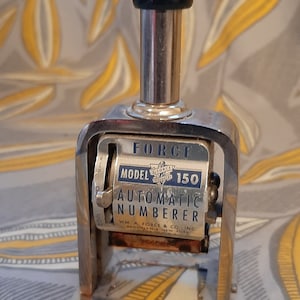 Puede incluir: Un numerador automático plateado con un mango negro. El numerador está etiquetado como "FORCE MODELO 150 NUMERADOR AUTOMÁTICO WM A FORCE & CO INC BROOKLYN N.Y."