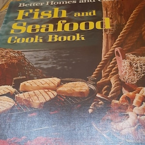 May include: A cookbook titled "Better Homes and Gardens Fish and Seafood Cookbook" with a red lobster on the cover. The book features a photo of grilled fish on a grill with a rope and a net in the background. The text on the cover reads "406 Great Tasty Recipes From Basic To Gourmet Ideas Using Fresh, Frozen And Canned Varieties".