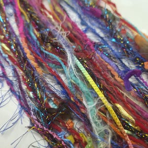 May include: A collection of multi-coloured art yarn strands featuring a variety of textures, including fuzzy, metallic, braided, and ribbon-like fibres in shades of pink, blue, purple, red, yellow, and green, arranged in a loose, vertical bundle.
