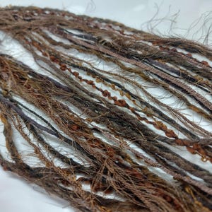May include: Close-up of brown and tan dreadlocks. The dreadlocks vary in thickness and texture, with some featuring decorative knots and braids. The colour palette includes shades of brown, tan, and a hint of purple, creating a natural, earthy aesthetic.