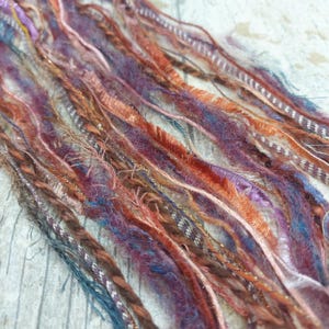 May include: A close-up of a collection of colourful yarn strands. The yarn features a mix of textures and shades, including orange, purple, blue, and brown. Some strands are fuzzy, others braided, and some have metallic threads. The arrangement is diagonal.