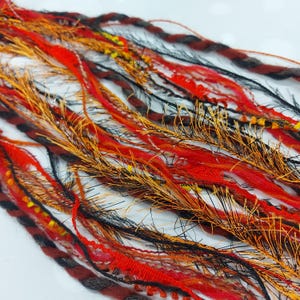 May include: Close-up of colourful strands of fibre in shades of red, orange, black, and brown. The fibres are of varying textures, including braided, fuzzy, and smooth. Some strands have yellow and silver accents. The fibres are likely for crafting or hair extensions.