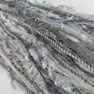 May include: A collection of assorted decorative yarns and ribbons in shades of grey, white, and muted blue. The strands feature varied textures including fuzzy, frayed, metallic, and braided elements, arranged in a parallel, flowing composition.