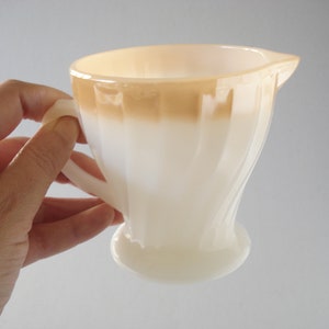 以下が含まれることがあります： 茶色のトップを持つ白いミルクガラスのクリーマー。クリーマーはフルーテッドデザインとハンドルが付いています。